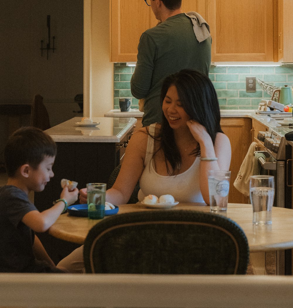 Zenia laughing at the kitchen table over breakfast with one of the kids, partner cooking in the background